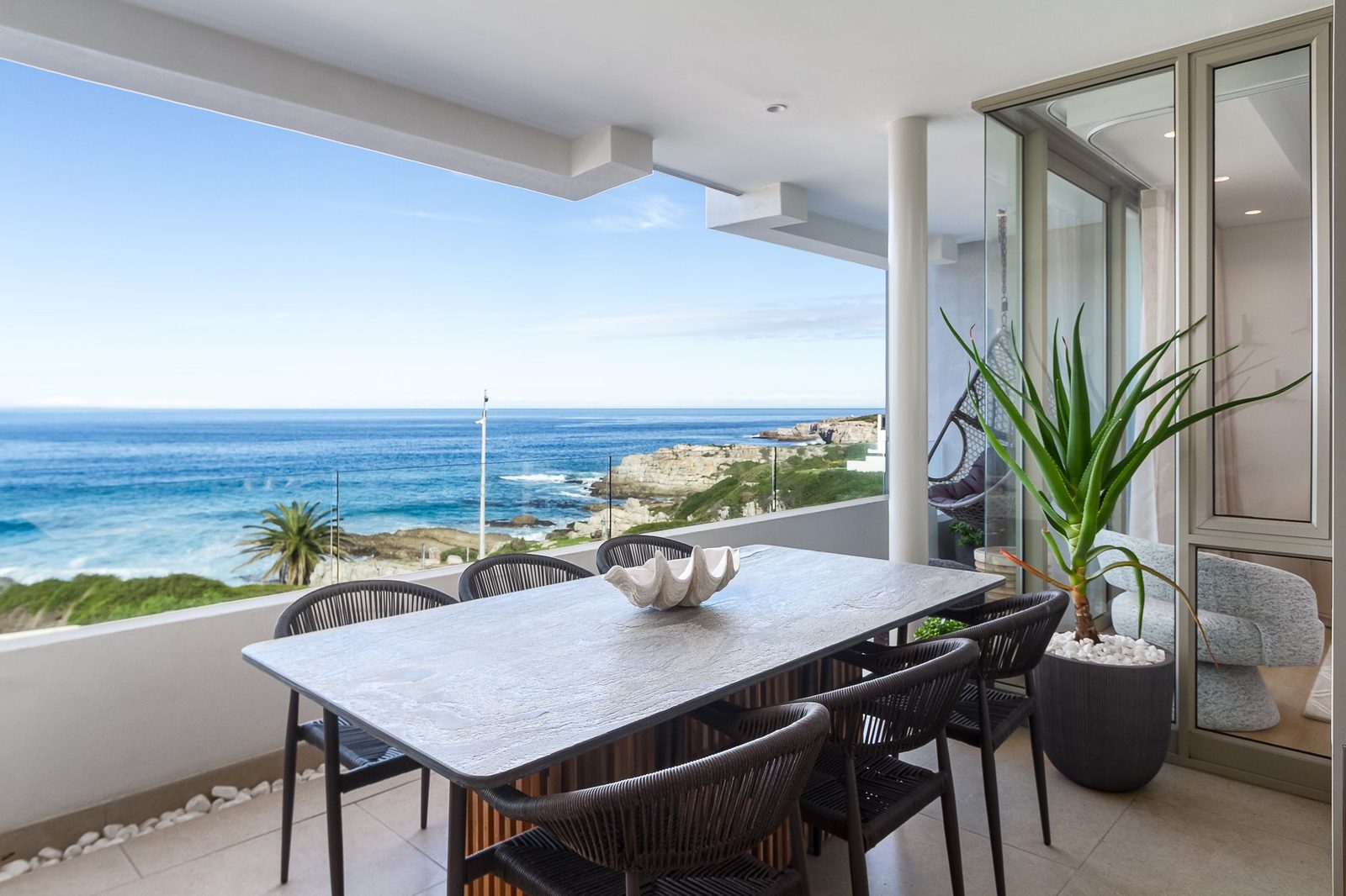 The Mira: covered balcony with a stone dining table and woven chairs above a rocky coastline