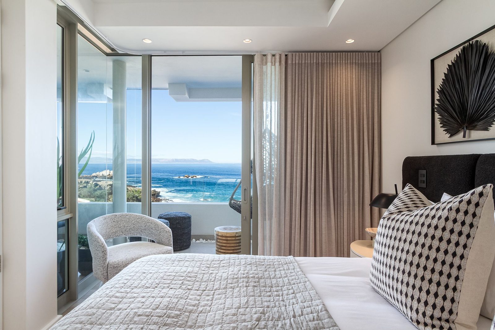 Bedroom opening onto a balcony above the sea, with linen curtains and a black palm frond artwork